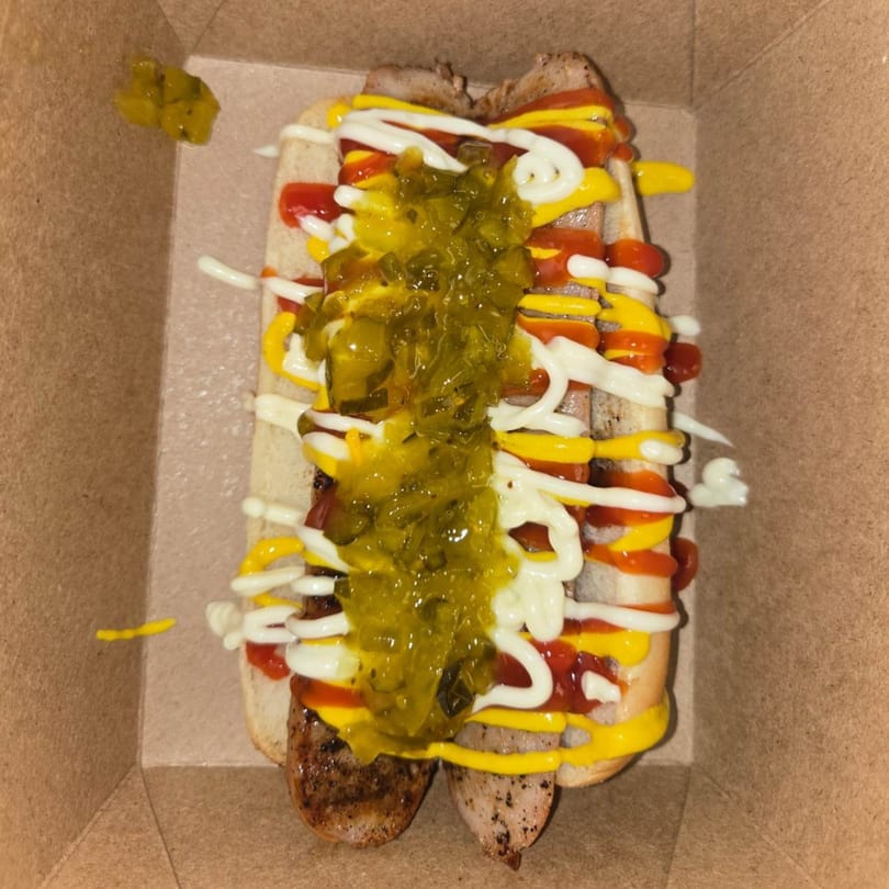 Two hot dogs in buns topped with relish, ketchup, mustard, and mayonnaise in a cardboard container.