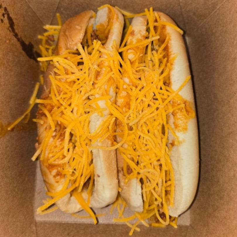 Two chili cheese hot dogs in a cardboard box, topped with shredded cheddar cheese.