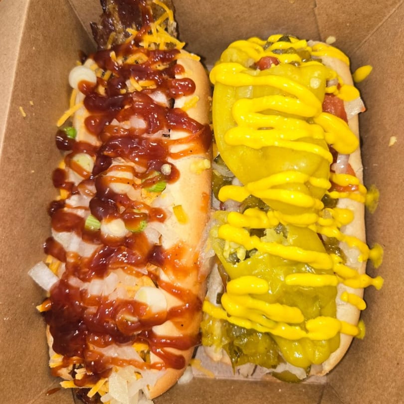 Two hot dogs in a cardboard tray, one with BBQ sauce and cheese, the other with relish and mustard.