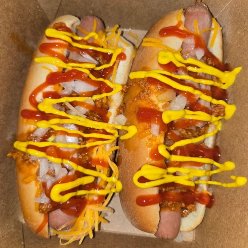 Two chili cheese hot dogs with diced onions, ketchup, and mustard in a cardboard box.
